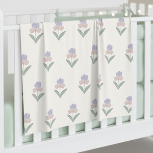Puede incluir: Una manta color crema con un patrón repetido de iris estilizados en tonos morado, rosa y verde. La manta está sobre una cuna blanca, mostrando el diseño floral. Los colores suaves y el patrón floral crean una estética relajante.