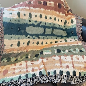 May include: A woven throw blanket with a colorful abstract pattern in shades of brown, green, and white. The blanket has a fringe border.
