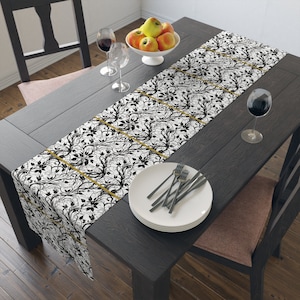 May include: A black and white table runner with a gold floral pattern. The runner is laid on a dark wood table with a bowl of apples, two wine glasses, and a plate with silverware.