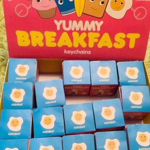 May include: A box of 20 blind boxes featuring a variety of breakfast-themed characters. The boxes are blue with a white egg design and the text "kidrobot" printed on the front. The box is labeled "YUMMY BREAKFAST keychains".