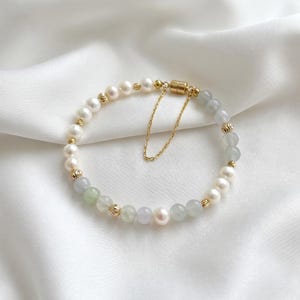 May include: A beaded bracelet featuring white freshwater pearls, translucent pale green jade beads, and small gold-toned metal spacers. The bracelet includes a gold-toned cylindrical magnetic clasp and a thin gold-toned safety chain attached.