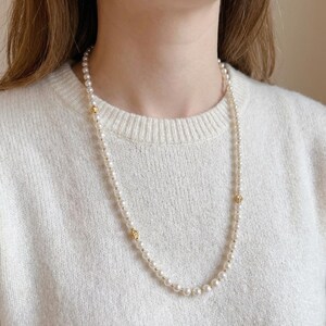 May include: A long pearl necklace with gold-coloured details. The necklace is made up of small, round, white pearls and has three decorative gold-coloured floral clasps. The necklace is worn over a white jumper.