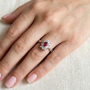 Puede incluir: Anillo de plata con un rub&iacute; central ovalado rodeado de piedras blancas m&aacute;s peque&ntilde;as. La banda del anillo tiene un dise&ntilde;o retorcido. El anillo est&aacute; en un dedo.