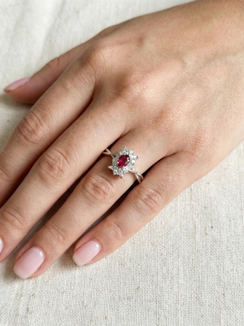 Puede incluir: Anillo de plata con un rub&iacute; central ovalado rodeado de piedras blancas m&aacute;s peque&ntilde;as. La banda del anillo tiene un dise&ntilde;o retorcido. El anillo est&aacute; en un dedo.
