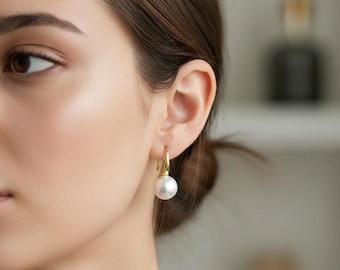 May include: A close-up of a gold-colored hoop earring with a large, white pearl. The earring is attached to an ear. The background is blurred, with neutral tones.