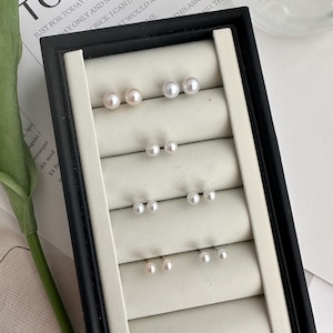 May include: A set of six pairs of pearl earrings displayed in a black velvet jewellery box. The earrings are arranged in three rows of two, with each pair featuring a small, white pearl.