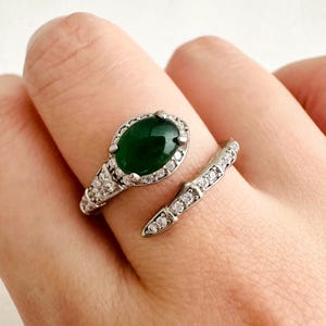 May include: A silver-toned bypass ring featuring a large, oval-shaped dark green stone set in a halo of small clear crystals. The band is open-ended and decorated with a series of small, sparkling clear crystals along both sides of the metal shank.