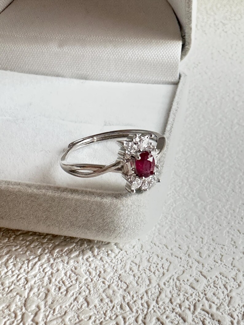 Puede incluir: Un anillo de plata con un rub&iacute; central de forma ovalada, rodeado de peque&ntilde;as piedras preciosas transparentes. La banda del anillo tiene un dise&ntilde;o delicado e entrelazado. El anillo se muestra en una caja de joyer&iacute;a gris, sobre una superficie blanca texturizada.
