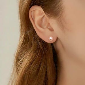May include: A close-up of a person's ear with a single, small, round, light pink pearl stud earring. The earring is centered on the earlobe. The person has long, light brown hair.