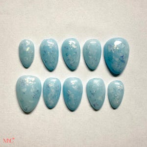 Puede incluir: Un juego de diez uñas postizas en color azul cielo claro con acabado brillante. Cada uña presenta escamas iridiscentes incrustadas que crean un efecto brillante y reflectante en la superficie. Las uñas tienen una forma de almendra redondeada.