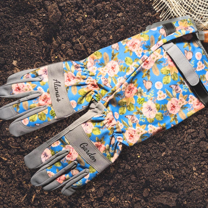 Peut inclure: Gants de jardinage bleus &agrave; motifs floraux avec des fleurs roses et jaunes. Les gants ont des accents gris et le texte "Alana's Garden" imprim&eacute; dessus. Les gants sont pos&eacute;s sur de la terre.