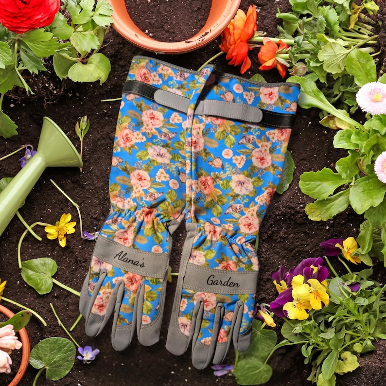 Peut inclure: Gants de jardinage bleus avec un motif floral de fleurs roses et p&ecirc;che. Les gants ont des accents gris et les mots "Alana's" et "Garden" imprim&eacute;s dessus. Les gants sont entour&eacute;s de terre, de fleurs et d'un arrosoir.