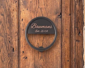 May include: A circular black metal door knocker featuring a horizontal bar with the engraved text "Baumans" and "Est. 2024" in a light-colored script font. The hardware is mounted on a vertical wooden door with a visible grain and natural texture.