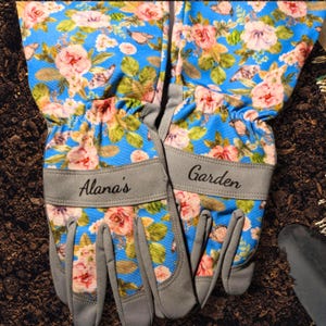 Peut inclure: Une paire de gants de jardinage avec un motif floral bleu, des roses roses et jaunes et des feuilles vertes. Les gants ont des poignets gris avec les textes "Alana's" et "Garden".