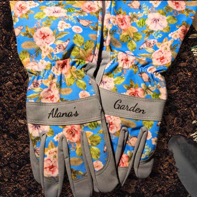 Peut inclure: Une paire de gants de jardinage avec un motif floral bleu, des roses roses et jaunes et des feuilles vertes. Les gants ont des poignets gris avec les textes "Alana's" et "Garden".
