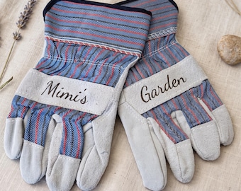 May include: A pair of gardening gloves featuring a blue, red, and black striped pattern on the back and light gray suede-like palms. The gloves have a light gray cuff section with the text Mimi's printed on one and Garden on the other.