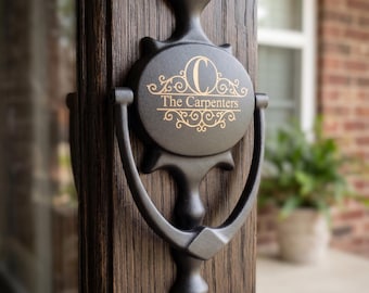 Peut inclure: Un heurtoir de porte en métal noir mat monté sur une porte en bois foncé. La plaque centrale circulaire présente une lettre C dorée en haut, le texte The Carpenters au milieu et des ornements en volutes au-dessus et en dessous du texte.