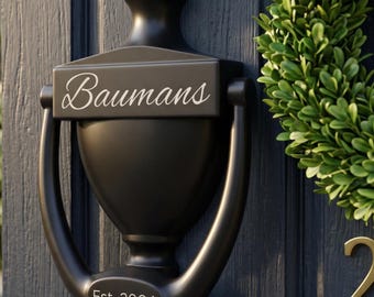 Peut inclure: Un heurtoir de porte en métal noir mat avec une forme d'urne classique. La barre horizontale supérieure présente le texte en écriture blanche "Baumans" et la section inférieure affiche le texte blanc "Est. 2004" sur une porte à panneaux gris foncé.