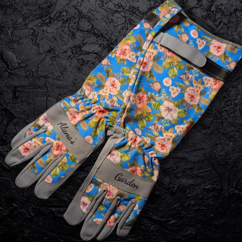 Peut inclure: Une paire de gants de jardinage bleus avec un motif floral de roses roses et de feuilles vertes. Les gants ont des paumes et des poignets en daim gris, avec le nom "Alana's" et le mot "Garden" imprim&eacute;s dessus. Les gants ont des sangles r&eacute;glables.