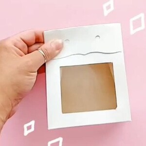 May include: White cardboard box with a window cut out. The box has two holes at the top and a wavy line across the top.