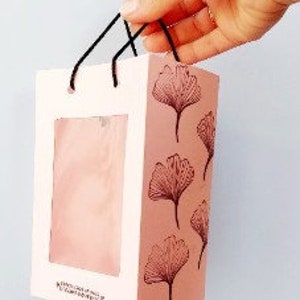 May include: A pink paper gift bag with a clear window and black handles. The bag has a ginkgo leaf design printed on it.