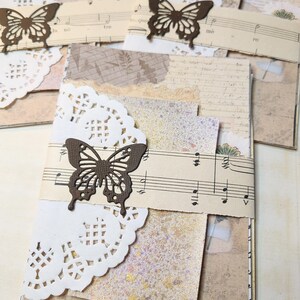Op de afbeelding: Handgemaakte papieren creaties met een vlinderthema. Elk stuk bevat gelaagd papier, vintage bladmuziek en decoratieve doilies. De vlinders zijn uit bruin karton gesneden en vormen een contrast met de zachte kleuren.