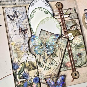 May include: A handmade scrapbook with a vintage aesthetic. Features include a map, floral illustrations, and decorative butterfly embellishments. The color palette includes shades of blue, green, and beige. The scrapbook is open, revealing multiple layers and compartments.