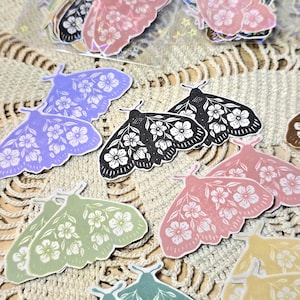 May include: A collection of moth-shaped stickers featuring floral patterns in various colors including purple, black, pink, green, and yellow. The stickers are displayed individually and in clear plastic sleeves, showcasing detailed white flower designs.
