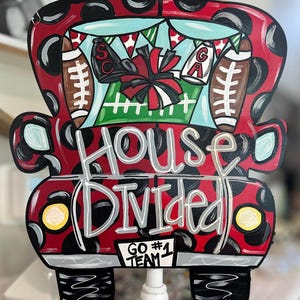 May include: A red and black wooden truck-shaped sign with the words "House Divided" in gray and white. The truck bed features a football field scene with footballs, a megaphone, and the letters "SC" and "GA". The sign reads "GO #1 TEAM".