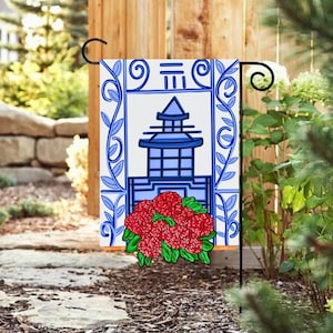Könnte beinhalten: Eine blaue und weiße Gartenflagge mit einem Pagoden-Design und roten Blumen. Die Flagge hat einen weißen Hintergrund mit einer blauen Pagode in der Mitte. Die Pagode hat ein blaues Dach und weiße Fenster. Die Flagge ist von einem blauen und weißen Rand mit einem floralen Design umgeben. Die Flagge hängt an einer schwarzen Metallstange.