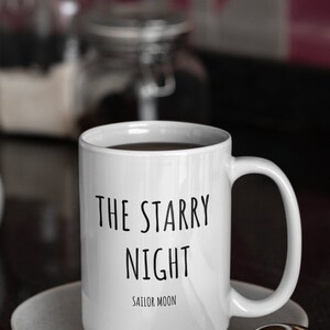 May include: White ceramic coffee mug with black text that reads "The Starry Night Sailor Moon". The mug is sitting on a white saucer.