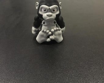May include: A black and white figurine of a gorilla sitting with its legs crossed. The gorilla has a serious expression on its face.