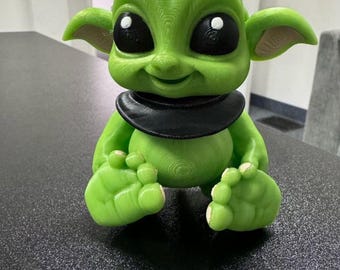 Puede incluir: Una figurita de alien verde con ojos grandes y un collar negro. La figurita está sentada con las piernas cruzadas y tiene una expresión amigable.