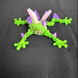 May include: A green frog toy with purple and pink accents. The frog has large, purple eyes and a pink, white, and purple design on its back. The frog's limbs are articulated and can be posed in different positions.