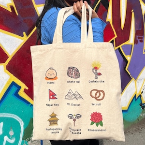 May include: A white tote bag with colorful illustrations of Nepalese cultural icons, including a momo, dhaka topi, dashain tika, Nepal flag, Mt. Everest, sel roti, Pashupatinath temple, birthplace of Buddha, and rhododendron.
