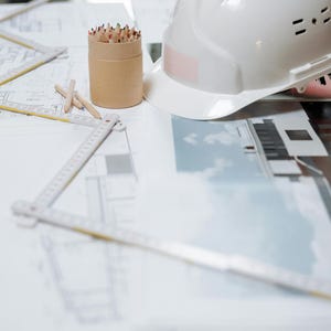 Könnte beinhalten: Ein weißer Bauhelm mit einem rosa Aufkleber liegt auf einem Tisch mit architektonischen Plänen. Ein zylindrischer Behälter enthält Buntstifte, und ein Zollstock und andere Werkzeuge sind ebenfalls vorhanden. Die Szene deutet auf ein Bau- oder Designprojekt hin.