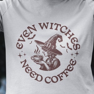以下が含まれることがあります： コーヒーを蒸したカップを持った魔女を描いた茶色の円形グラフィックが施された白いTシャツ。グラフィックの周りに「Even Witches Need Coffee」というテキストが書かれています。