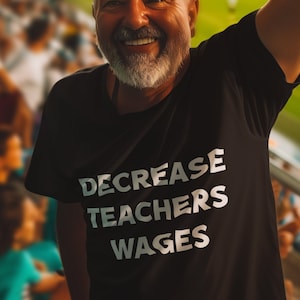 Puede incluir: Un hombre con una camiseta negra con el texto "DECREASE TEACHERS WAGES" impreso en blanco. Está sonriendo y levantando su mano derecha en el aire. Está de pie en un estadio con una multitud de personas en el fondo.