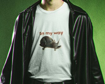 Puede incluir: Un hombre con una chaqueta de cuero negra y pantalones negros, sosteniendo una bolsa negra y una pistola. Lleva una camiseta blanca con el texto "On my way" y un gráfico de un caracol.