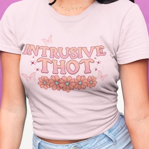 May include: A white t-shirt with a pink and orange graphic that says "Intrusive Thot" with butterflies and flowers around the text. The model is wearing the shirt and blue denim shorts.