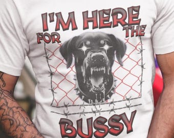 Könnte beinhalten: Weißes T-Shirt mit dem Text "I'M HERE FOR THE BUSSY" in Rot und Schwarz. Die Grafik zeigt einen knurrenden Hund hinter Stacheldraht. Die Person lächelt und trägt eine Uhr und ein Armband.