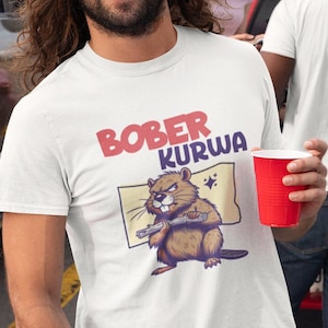 Op de afbeelding: Een man draagt een wit T-shirt met een beverafbeelding en de tekst "BOBER KURWA" en grilt worstjes en hamburgers op een zwarte houtskoolgrill. Hij houdt een rood kopje en een tang vast. Er ligt een kolf maïs op de grill.