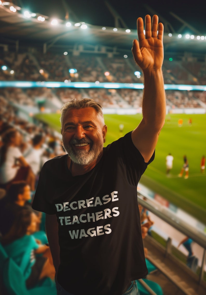 May include: A man wearing a black t-shirt with the text "DECREASE TEACHERS WAGES" printed in white. He is smiling and raising his right hand in the air. He is standing in a stadium with a crowd of people in the background.