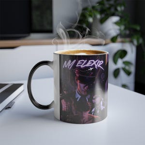 May include: A colour-changing coffee mug with a black handle and rim. The mug features a design with the text "MM Elixir" and an illustration of a person in a hat. The mug is filled with a hot beverage, with steam rising from the top.