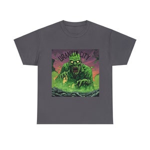 以下が含まれることがあります： 暗いグレーのTシャツに、緑色のモンスターのグラフィックと黒い文字で「Uranium City」と書かれています。