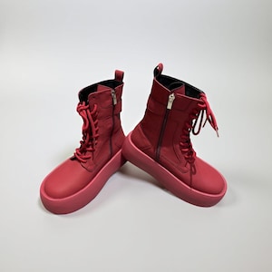 May include: A pair of red combat boots with thick soles. The boots have laces, zippers, and a high-top design. The boots are made of a smooth material and are set against a white background.