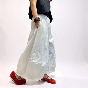 Op de afbeelding: Wijde witte broek met een geplooide elastische tailleband, zijzakken en een geplooide zoom. De broek heeft een volumineus silhouet en wordt gecombineerd met felrode platte schoenen met een strikdetail op de voorkant van elke schoen.