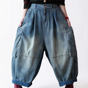May include: High-waisted blue denim pants featuring a loose, wide-leg silhouette with pleated details at the waist, large side cargo pockets, and a faded wash finish. The pants have a button closure and a wide hem at the bottom of each leg.