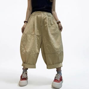 Puede incluir: Pantalones cortos de color beige con corte holgado, cintura alta, cierre de botón y cremallera, y costuras horizontales a la altura de los muslos. Los pantalones presentan una silueta ancha con dobladillos vueltos en la parte inferior.