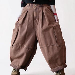 May include: Brown cargo pants featuring a high-waisted design with front pleats, side cargo pockets, and a loose, wide-leg silhouette. The pants have a button closure, a zip fly, and cuffed hems, providing a relaxed fit with visible stitching.